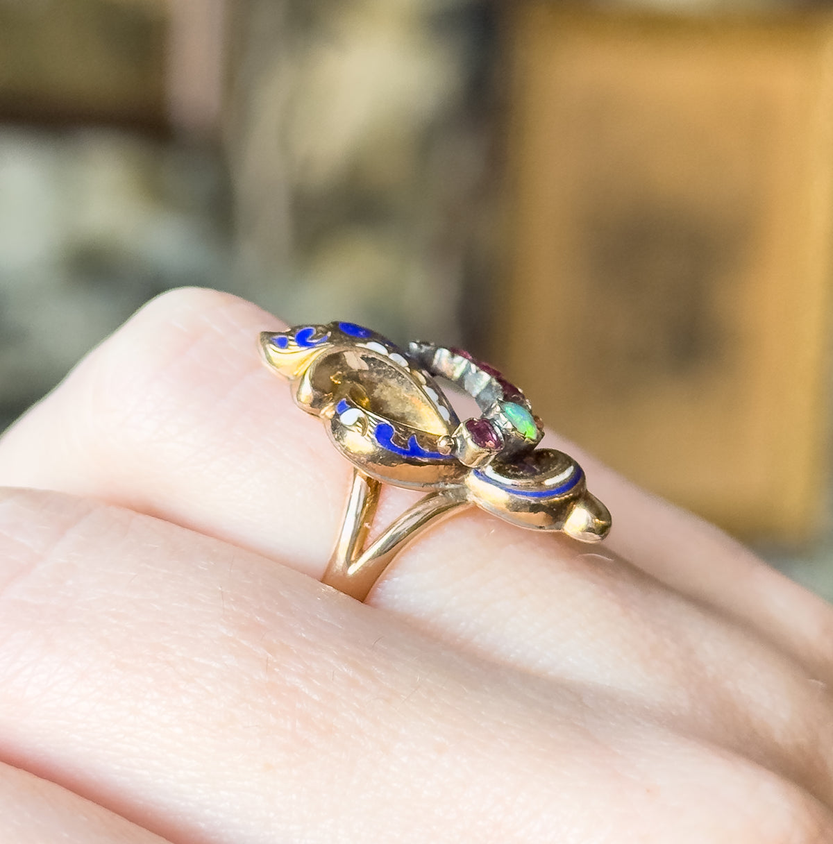 Shown on a finger from a low side angle, the polished yellow gold shank rising from the band to the raised face, with cobalt blue enamel along its edge and the opal and rubies at the far end.