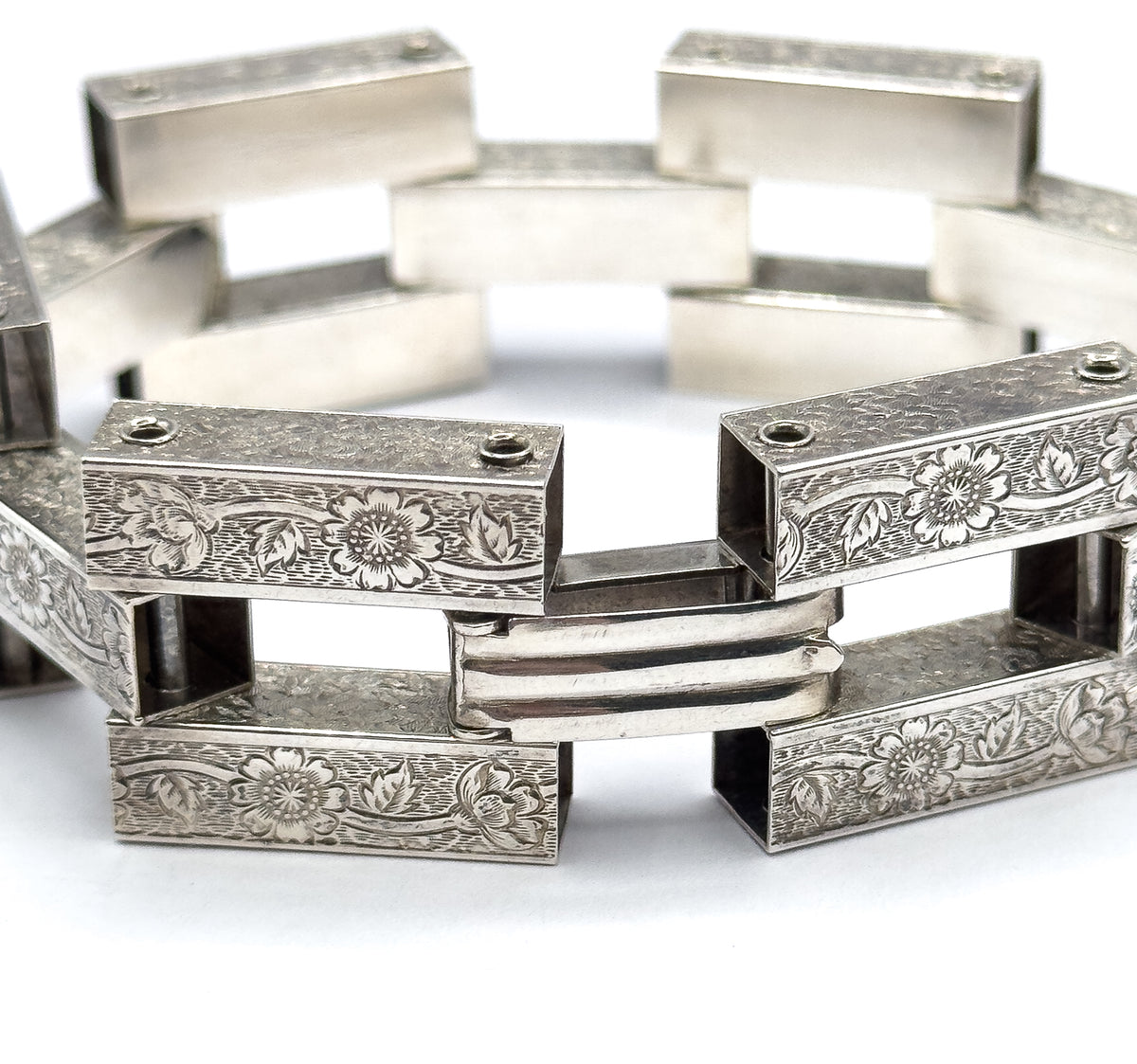 Tight view of the point where two engraved links meet, with a smooth ribbed connector bar bridging them and the rest of the bracelet curving away blurred behind.