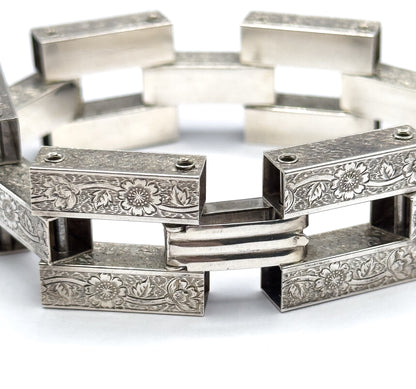 Tight view of the point where two engraved links meet, with a smooth ribbed connector bar bridging them and the rest of the bracelet curving away blurred behind.