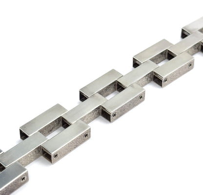 Close raking view along the links, where light catches the smooth flat tops of the bars while the floral texturing shows on their sides, with a hinge pin visible at the end of each bar.