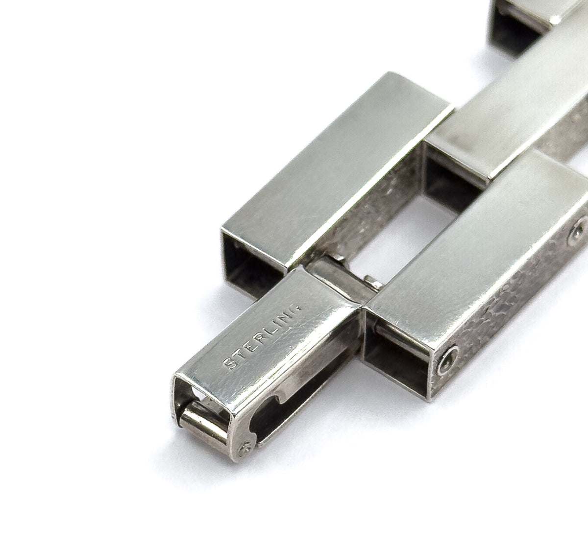 Tight close-up of the clasp end, where a slim tongue bar slides into the hollow square opening of a link, with small pins and hinge points visible between the broad bars.