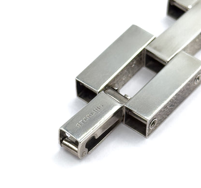 Tight close-up of the clasp end, where a slim tongue bar slides into the hollow square opening of a link, with small pins and hinge points visible between the broad bars.