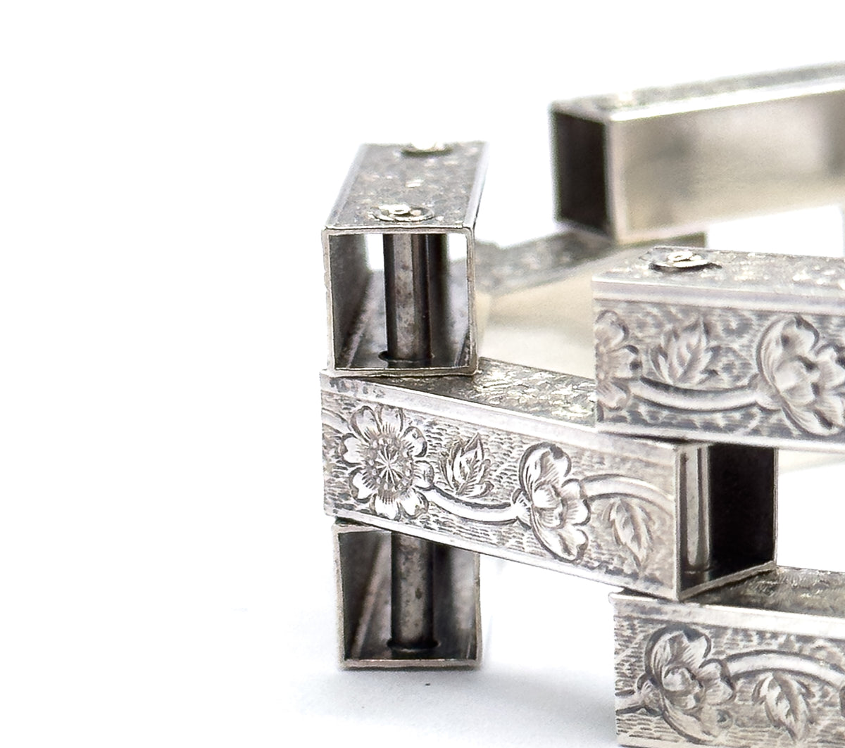 Extreme close-up of the gate links stacked at angles, showing engraved flowers and leaves across the side faces, hinge pin heads on the flat tops, and the hollow square bar ends.