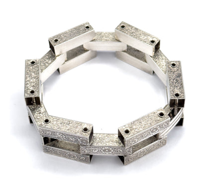 Tilted three-quarter view of the standing ring, where the near links lean toward the camera so their floral-engraved outer faces and the pierced square openings between bars are both visible.