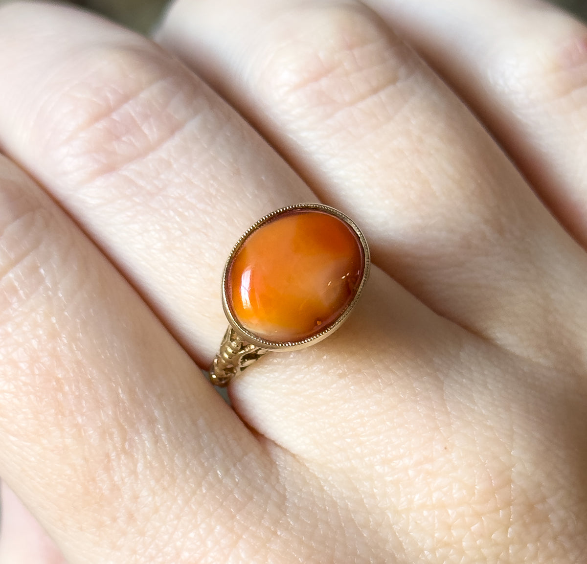 Close overhead view on the finger, filling the frame with the oval coral cabochon's warm orange tones and paler translucent patches inside its beaded bezel.