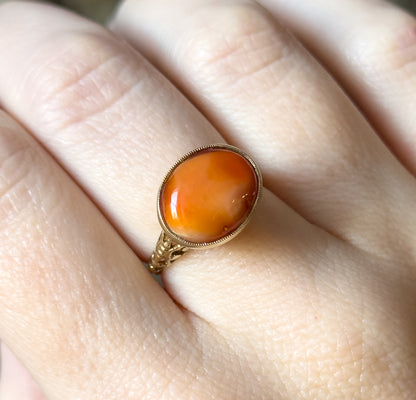 Close overhead view on the finger, filling the frame with the oval coral cabochon's warm orange tones and paler translucent patches inside its beaded bezel.