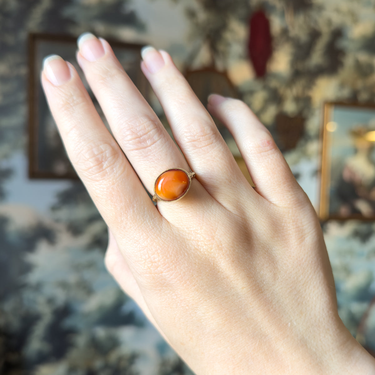 The whole hand shown against a soft painted backdrop, the ring on the ring finger giving a sense of its modest scale beside the knuckle.