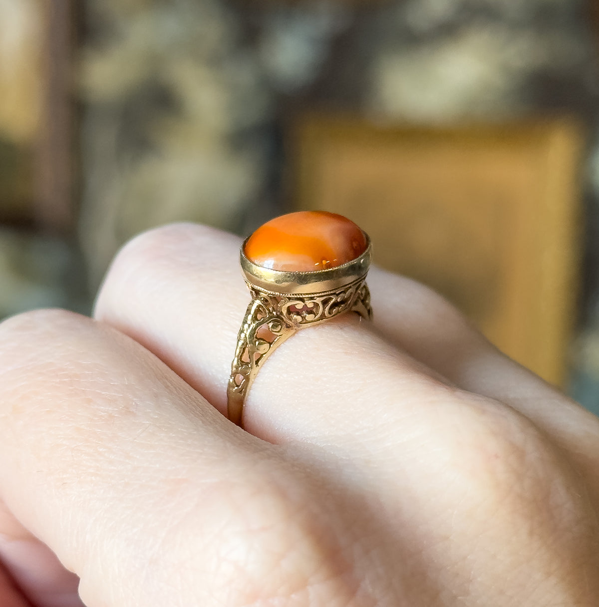 Art Deco coral cabochon filigree ring in 14K yellow gold, shown on a finger, the oval stone in a milgrain bezel above pierced filigree shoulders tapering to a slim shank.