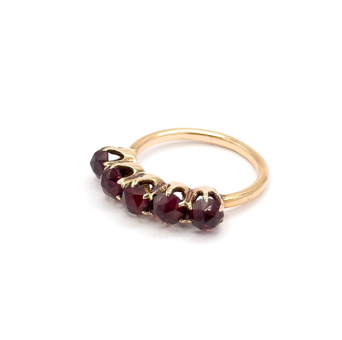Low three-quarter angle from the end of the row, the five garnets receding one behind another in their clawed yellow gold prongs while the plain round shank arcs up and away behind.