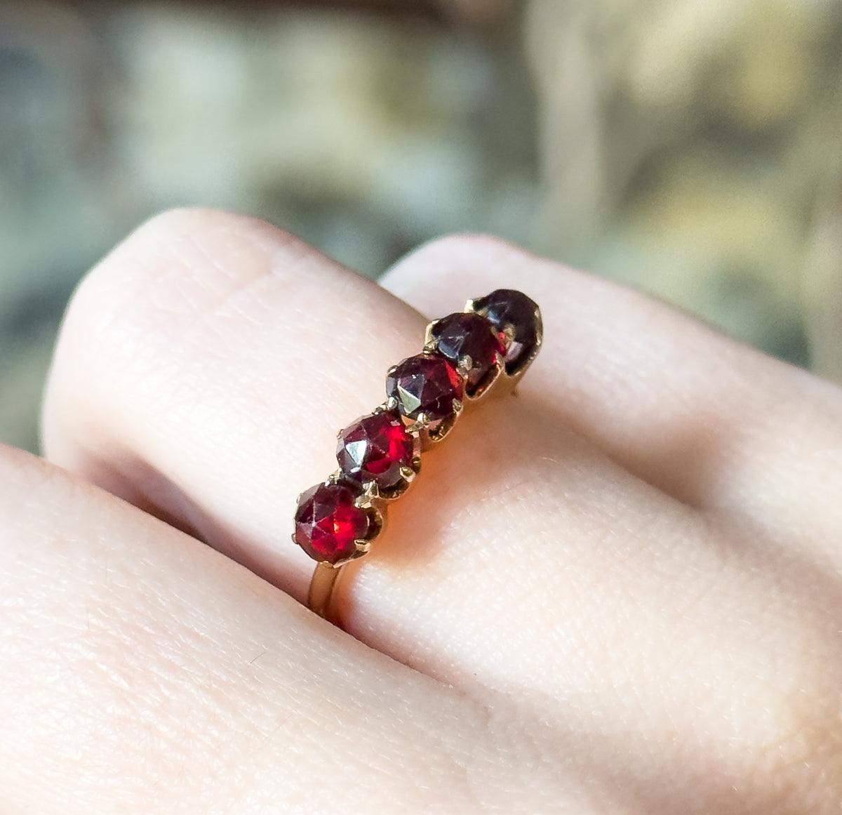 Macro close-up shown on a finger, the row of five deep red stones catching light at an angle so the individual prongs and the narrow yellow gold shank curving beneath are clearly visible.