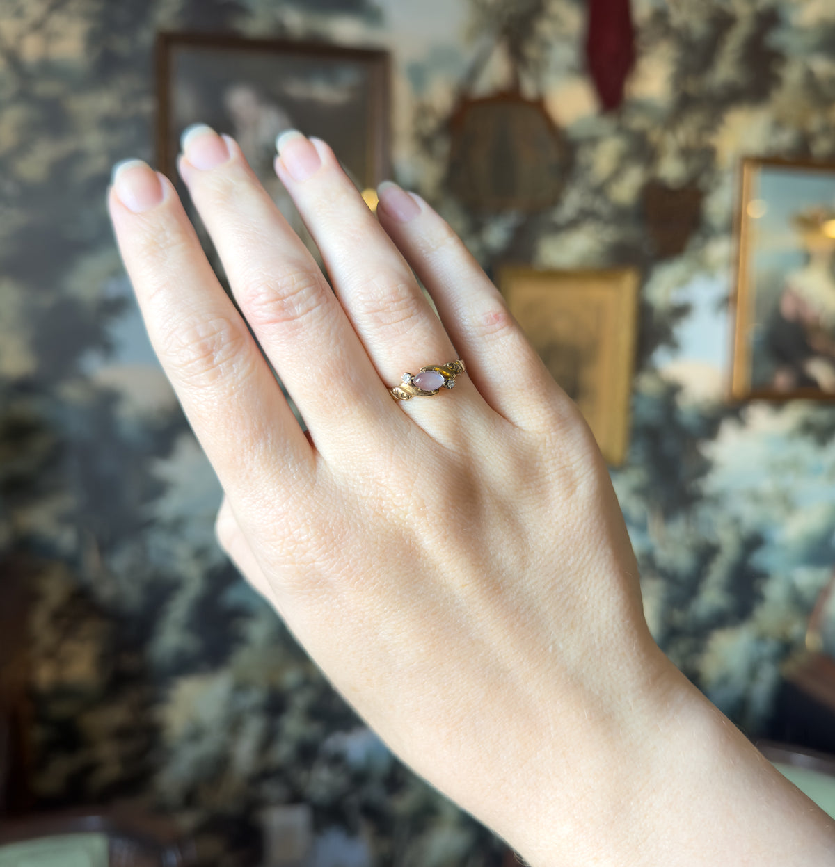 Shown on the ring finger of a raised hand against a soft painted backdrop, the ring reading as a small diagonal glint of yellow gold with its pale cabochon at this distance.