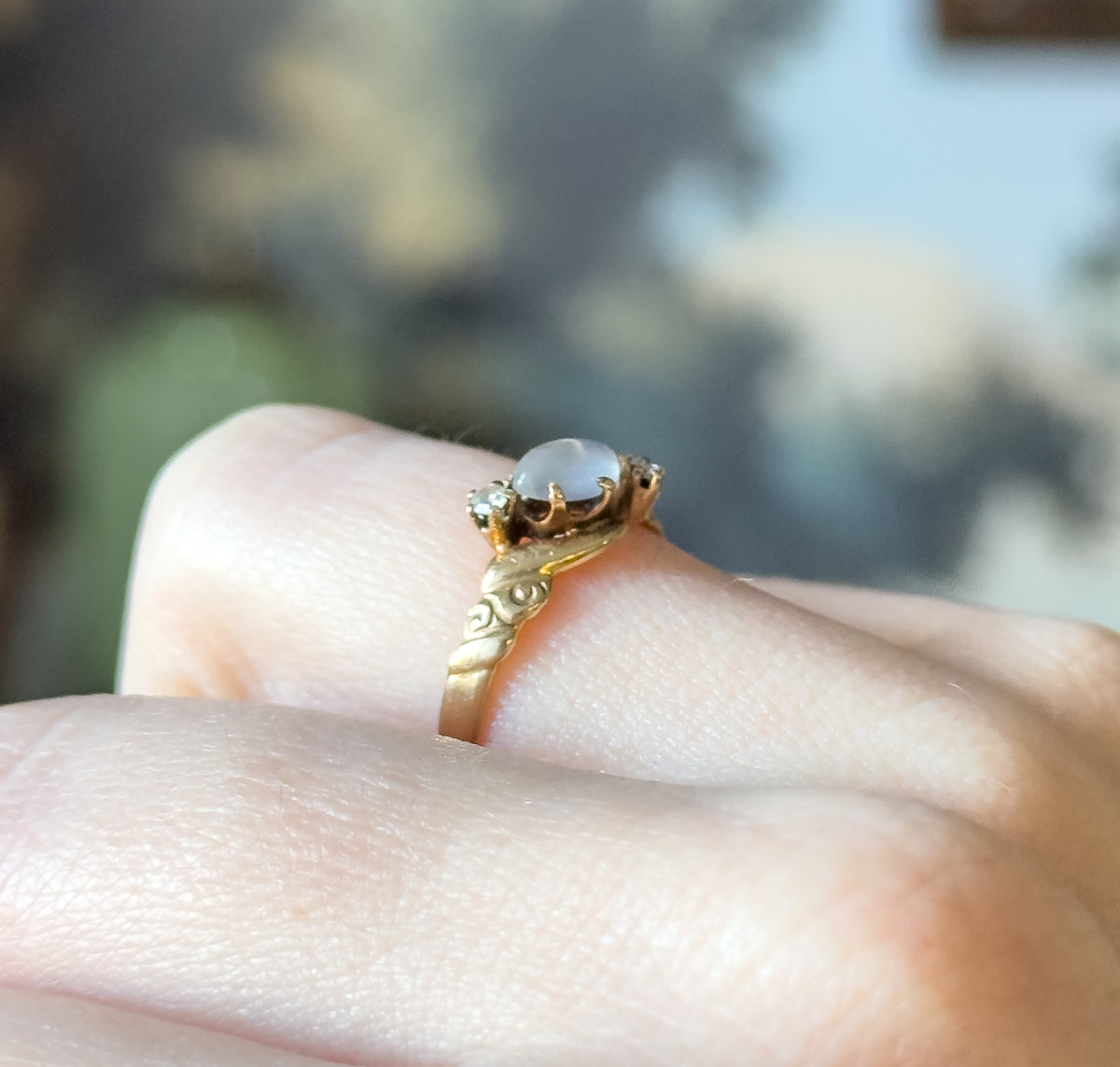 Victorian moonstone and diamond ring in 14K yellow gold, shown on a finger in close-up, the prong-set moonstone cabochon flanked by two small diamonds above a swirled shank.