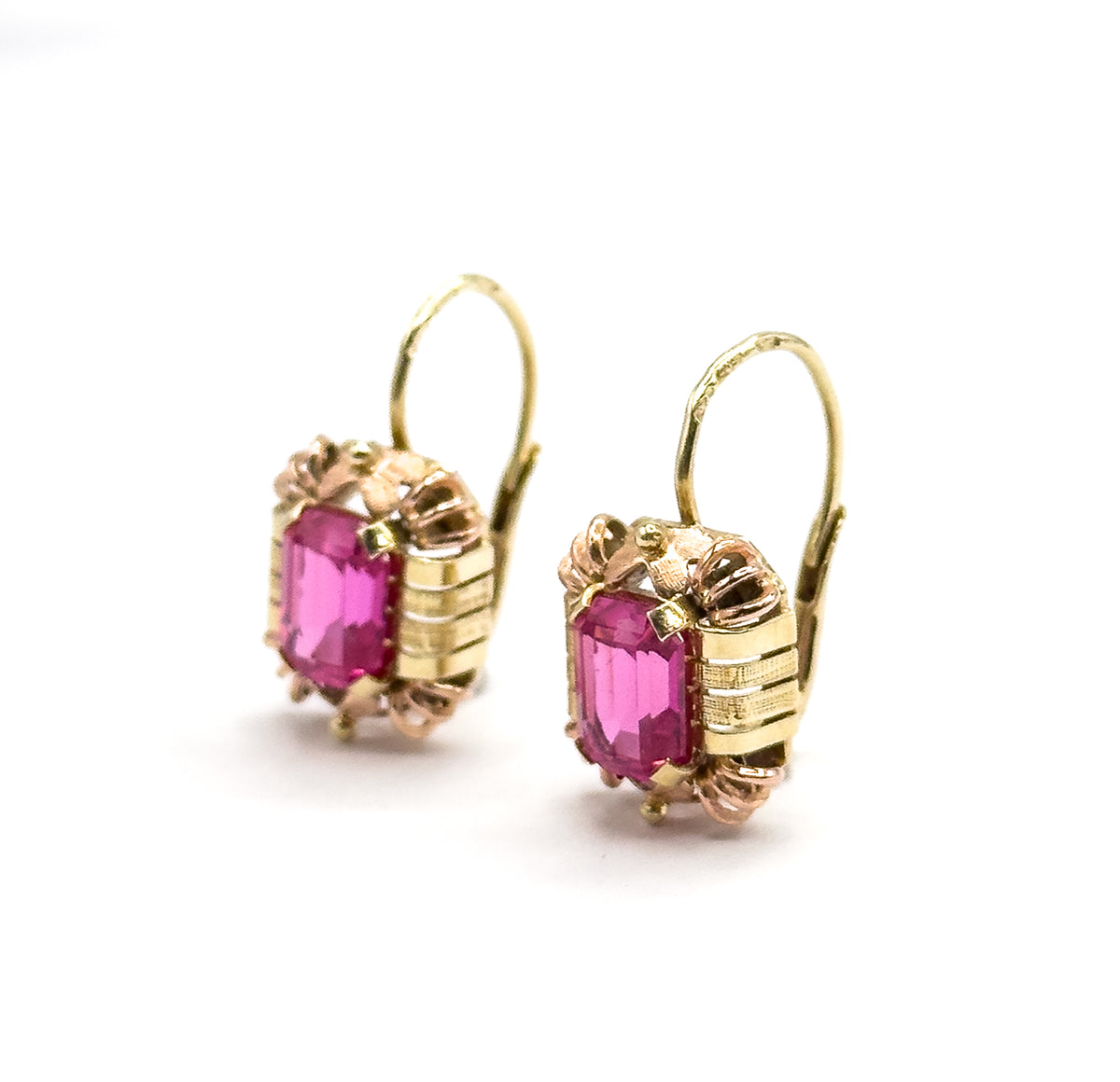 Close three-quarter view from the side, the ribbed yellow bars running vertically down each frame and the rose-gold prongs reaching across the emerald-cut rubies, hooks curving up behind.