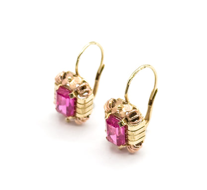 Overhead three-quarter view with the pair set diagonally across the frame, hooks closed and arcing up to the right, ribbed yellow bars and rose-gold prongs framing each emerald-cut ruby.