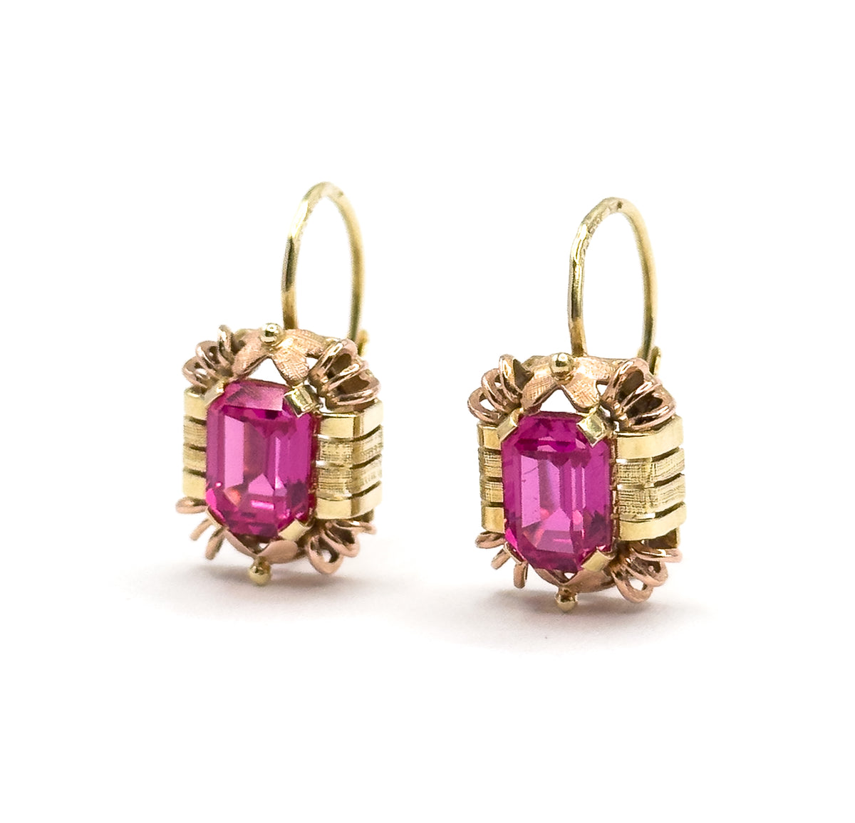 Both earrings standing upright side by side and facing forward, hooks open and looping above each frame, the emerald-cut rubies squared between ribbed yellow bars and rose-gold prongs.
