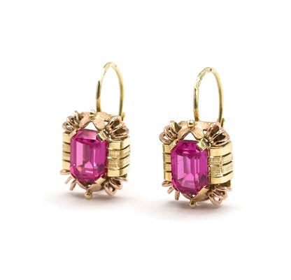 Both earrings standing upright side by side and facing forward, hooks open and looping above each frame, the emerald-cut rubies squared between ribbed yellow bars and rose-gold prongs.