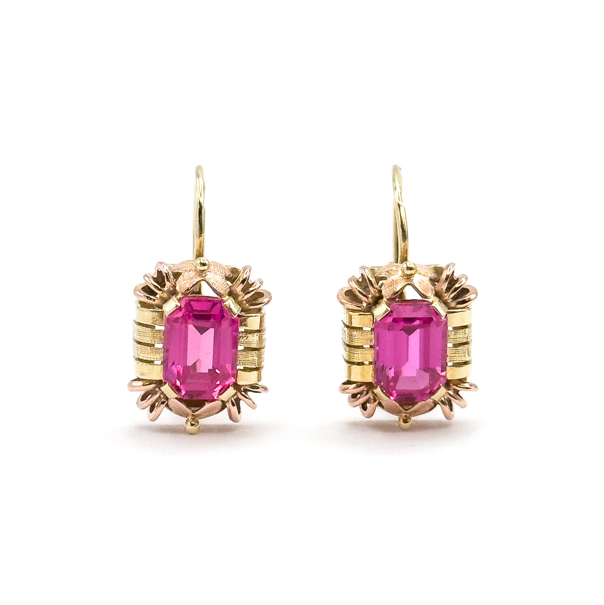 Straight-on front view of the pair, each emerald-cut ruby framed by ribbed yellow bars with rose-gold claws above and below and a small bead at the base, hooks rising in tall open curves.