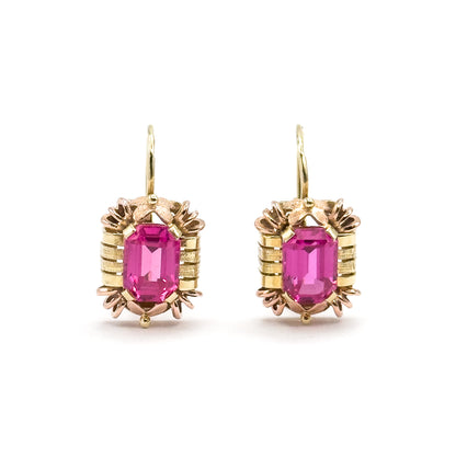 Straight-on front view of the pair, each emerald-cut ruby framed by ribbed yellow bars with rose-gold claws above and below and a small bead at the base, hooks rising in tall open curves.