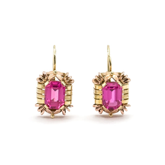 Straight-on front view of the pair, each emerald-cut ruby framed by ribbed yellow bars with rose-gold claws above and below and a small bead at the base, hooks rising in tall open curves.