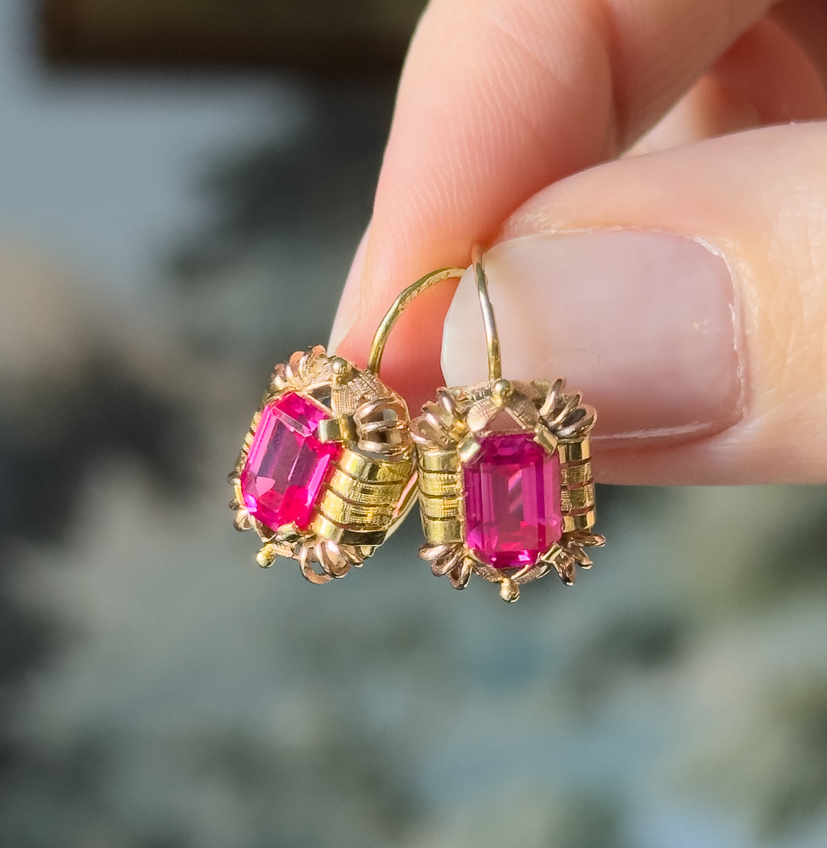 Retro lab-created ruby drop earrings in 14K two-tone gold, held between finger and thumb, the deep pink emerald-cut stones prong-set in textured yellow and rose frames with hook fittings.