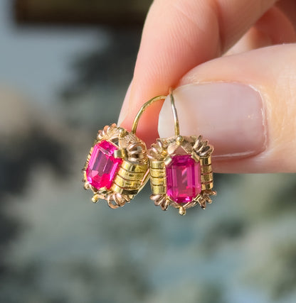 Retro lab-created ruby drop earrings in 14K two-tone gold, held between finger and thumb, the deep pink emerald-cut stones prong-set in textured yellow and rose frames with hook fittings.