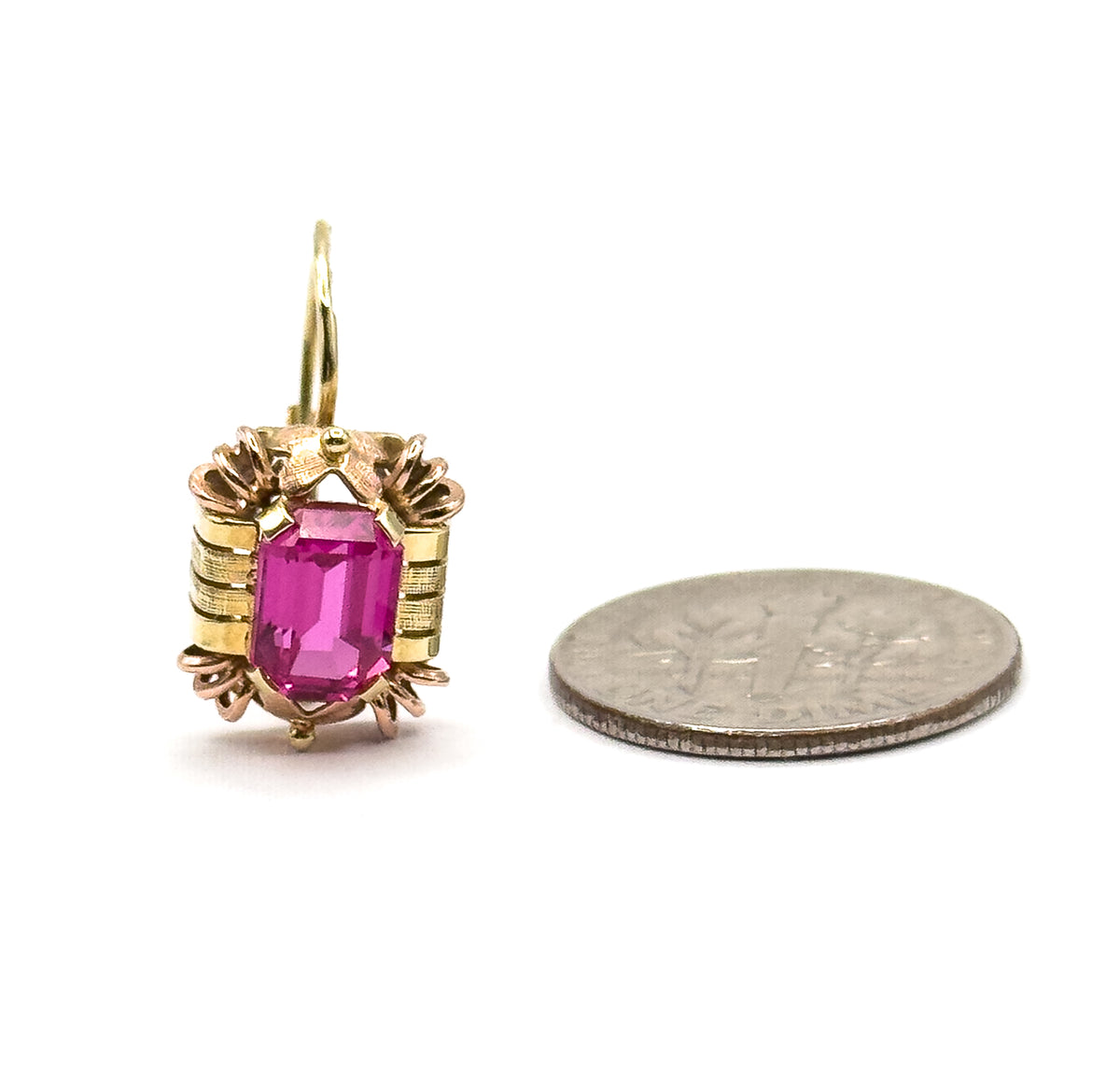 One earring beside a coin for scale, the deep pink emerald-cut ruby prong-set in its textured two-tone frame with the hook curving above, the stone roughly a third of the coin's width.