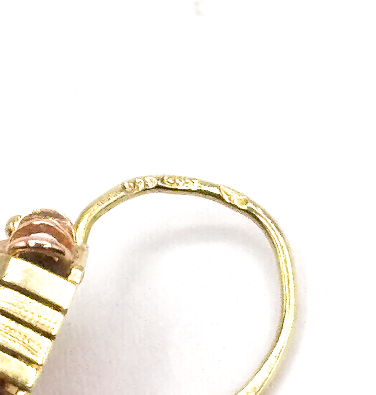 Extreme close-up of the curved hook wire, where tiny stamped marks are struck into the metal, with the rose-gold prongs and textured yellow frame at the lower left edge.