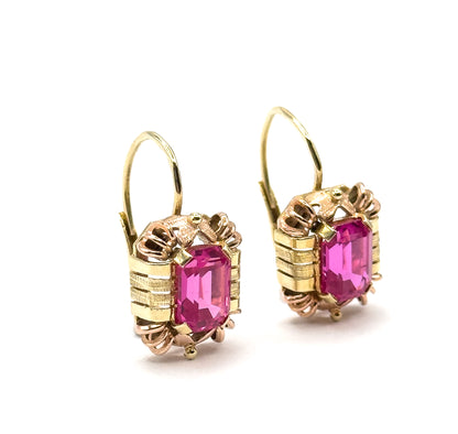 Three-quarter front view of both earrings tilted towards the viewer, the emerald-cut rubies bright between ribbed yellow bars and rose-gold prongs, hooks arcing up behind.