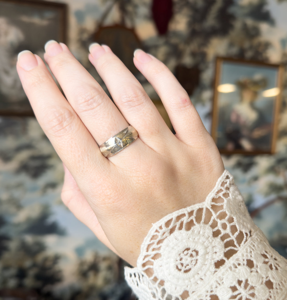The hand is shown raised against a soft floral backdrop, the wide band on the ring finger catching light across its face so the small diamond sits bright at the centre.
