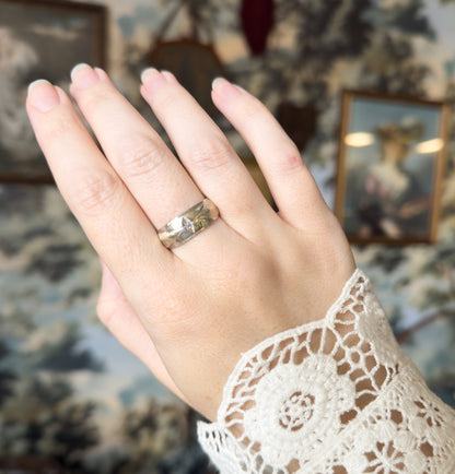 The hand is shown raised against a soft floral backdrop, the wide band on the ring finger catching light across its face so the small diamond sits bright at the centre.