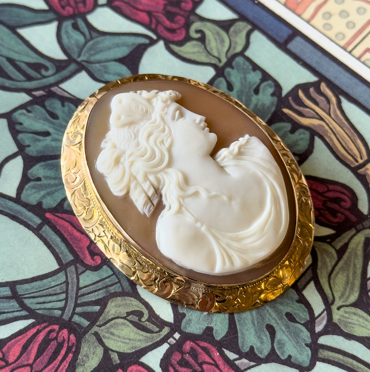 The brooch laid face up on a leafy patterned surface, the whole oval seen from above so the raised profile and the engraved floral border of the gold mounting read clearly.
