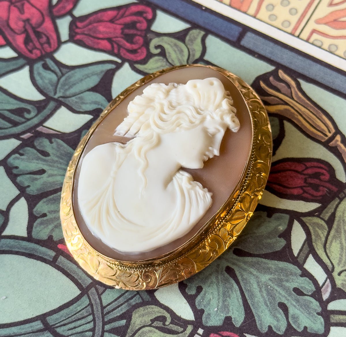 Seen at an angle on the same floral pattern, low enough to show the depth of the carved relief and the engraved side wall of the gold mounting rising around the shell.