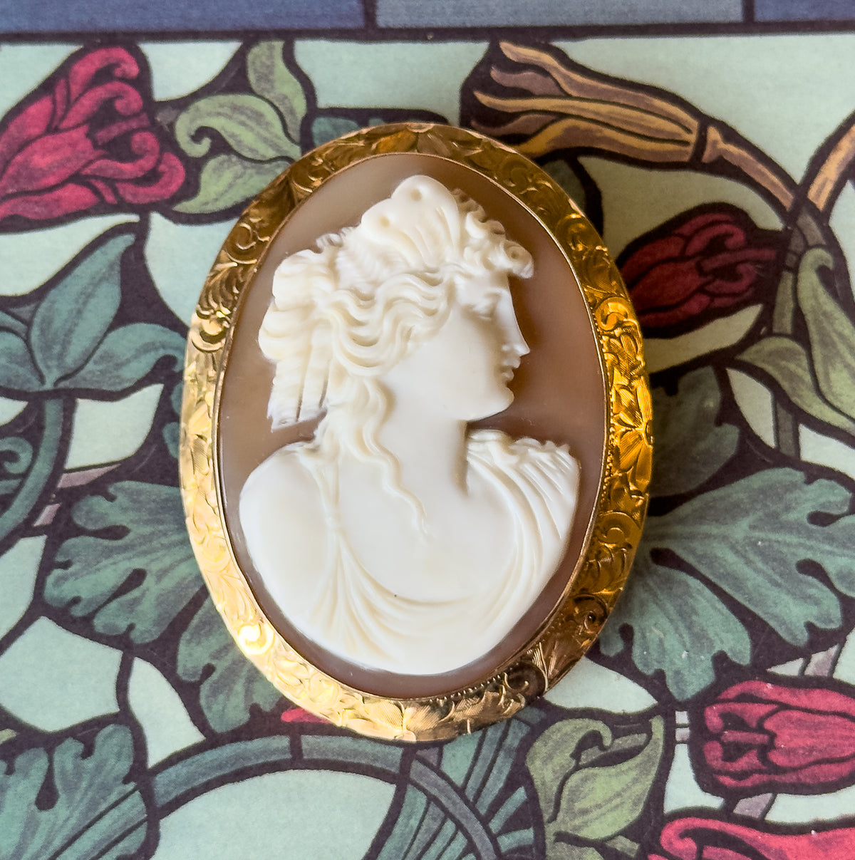 Close crop on the face-up brooch against the leafy pattern, filling the frame so the carved curls, the profile and the folds of drapery in creamy white against caramel brown show in detail.