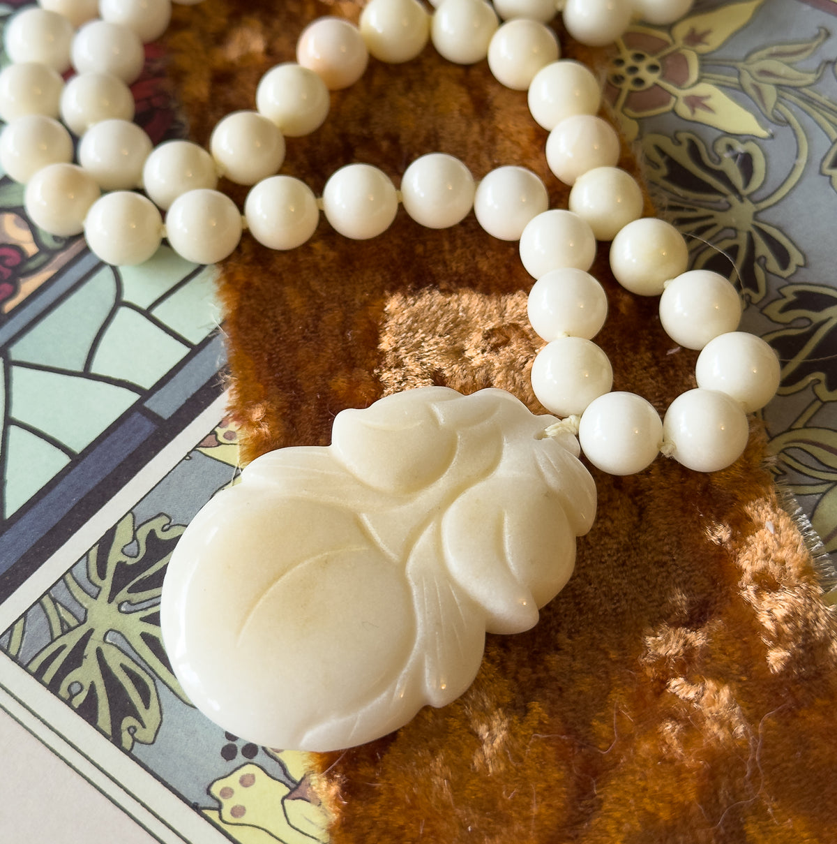 The strand loops across rust-coloured velvet and a decorative printed page, the carved gourd pendant at lower left, with one bead near the top showing a soft peach tint among the creamy white.
