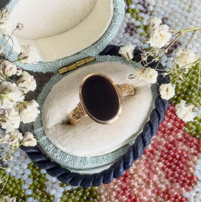 Now resting face-up in a pale open ring box among dried white flowers, the oval black agate filling its smooth bezel with engraved shoulders curving away on each side.