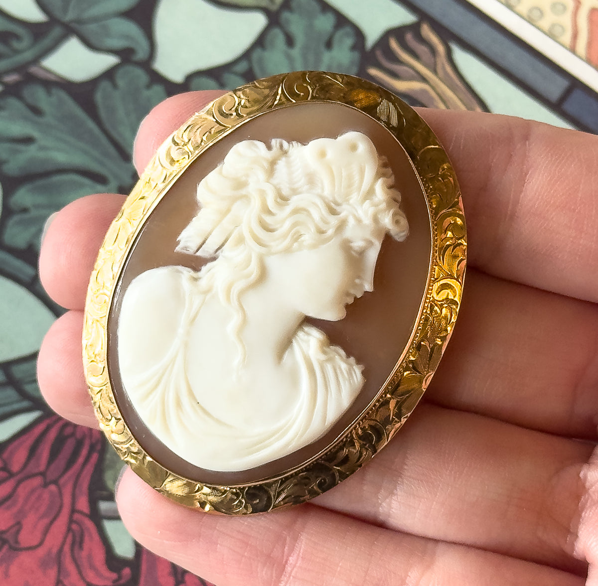 Almost the same view with slightly different angle, tilted closer so the engraved floral scrollwork on the gold oval and the carved curls and drapery of the profile stand out against the fingers.