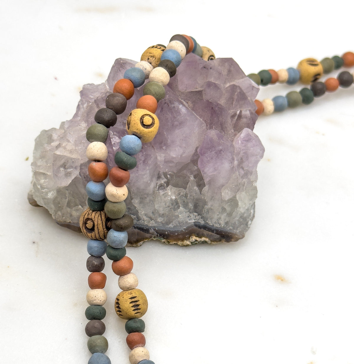 Strands draped over a chunky pale purple rock on white, with the beads trailing down in front of it and two of the larger marked ochre beads facing the camera.