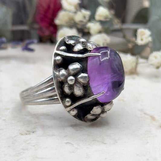 Silver ring with a purple gemstone on a textured surface with flowers in the background