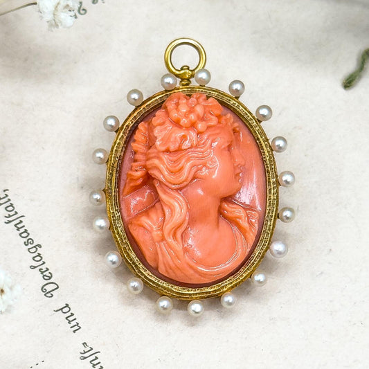 Coral cameo pendant with gold frame and pearl accents on a textured surface with floral elements.