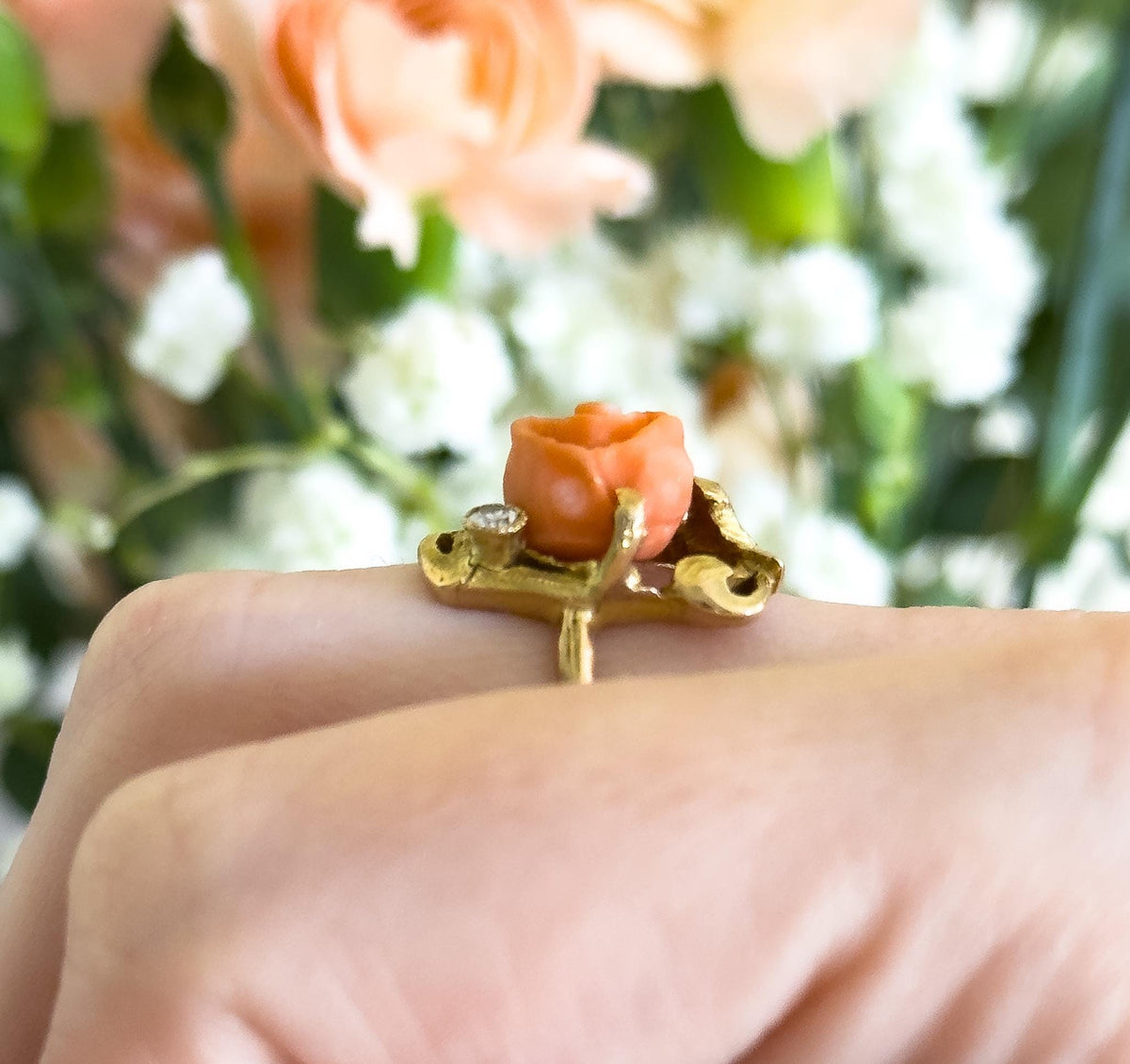 Close side-on view at knuckle level, shown on a finger, so the gold gallery's scrolled openwork and the shank beneath the coral rose and small bezel-set diamond are visible against blurred peach and white blooms.