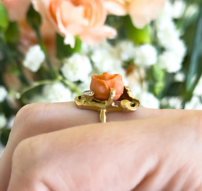 Close side-on view at knuckle level, shown on a finger, so the gold gallery's scrolled openwork and the shank beneath the coral rose and small bezel-set diamond are visible against blurred peach and white blooms.