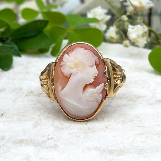 Vintage-style cameo ring with gold setting on a white surface with greenery and flowers in the background