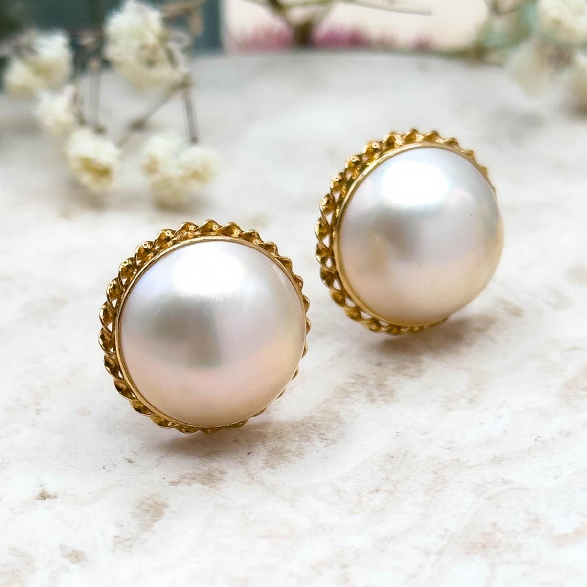 Pearl earrings with gold accents on a light surface with blurred flowers in the background