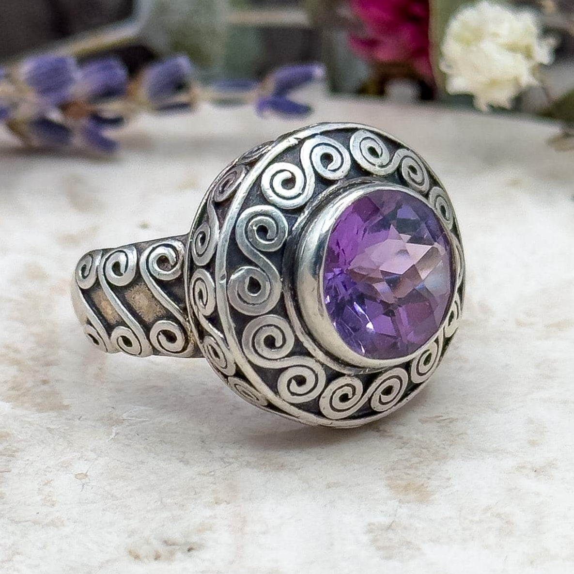 Silver ring with a purple gemstone on a textured surface with flowers in the background