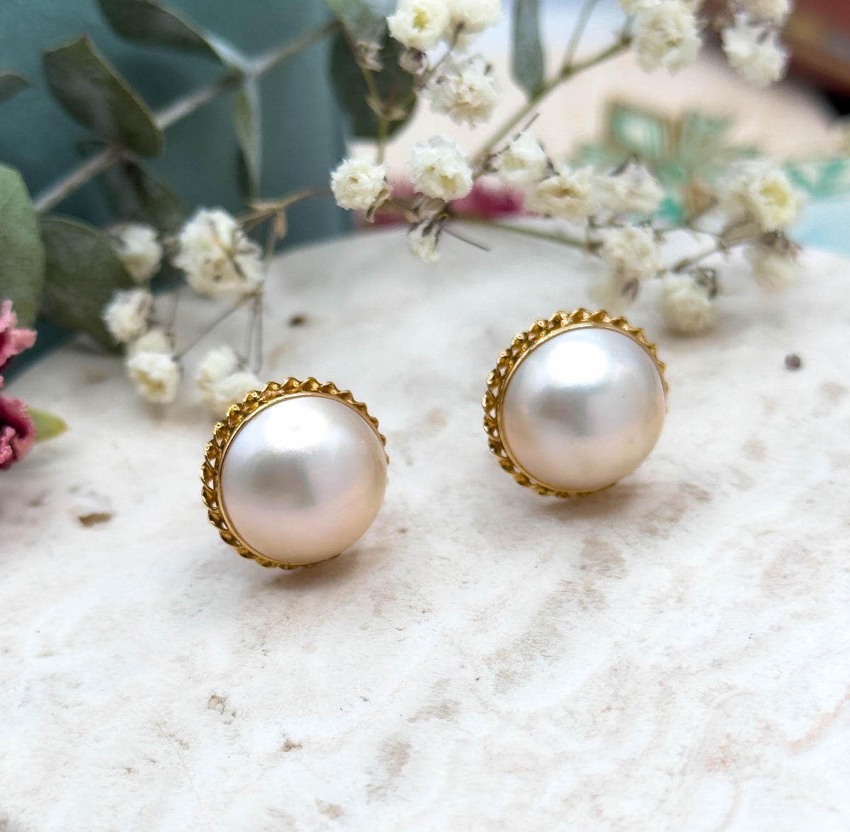 Close front view of the pair angled side by side on a pale stone slab with dried baby's breath and eucalyptus behind, showing the rope twist yellow gold bezel circling each domed pearl.