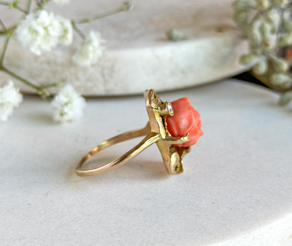 Almost the same view with slightly different angle, the ring tilted so the flat rectangular face and the gold scrollwork behind the coral rose come forward above the thin shank on the pale stone.
