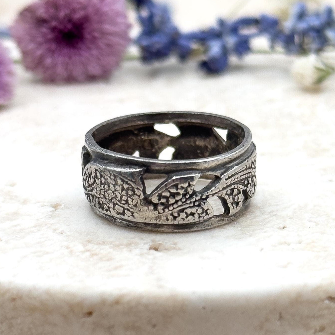 Silver ring with intricate design on a marble surface with flowers in the background