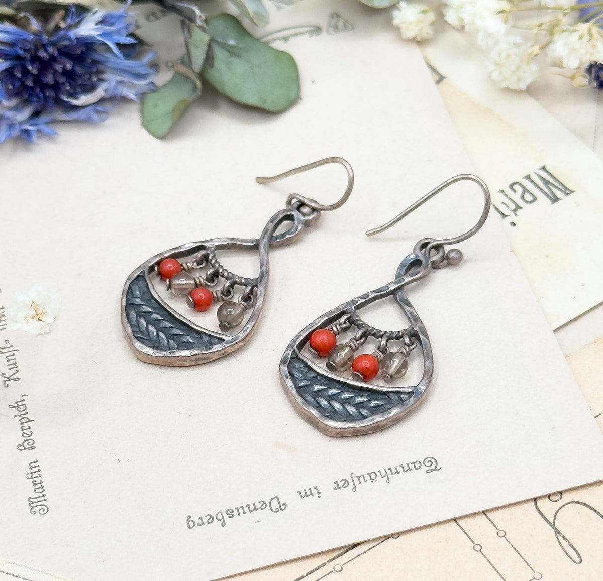 Closer crop on the two teardrops, showing the dark patina in the braided panels and the small linked wires holding each red coral and clear glass bead, with dried flowers on the printed card behind.
