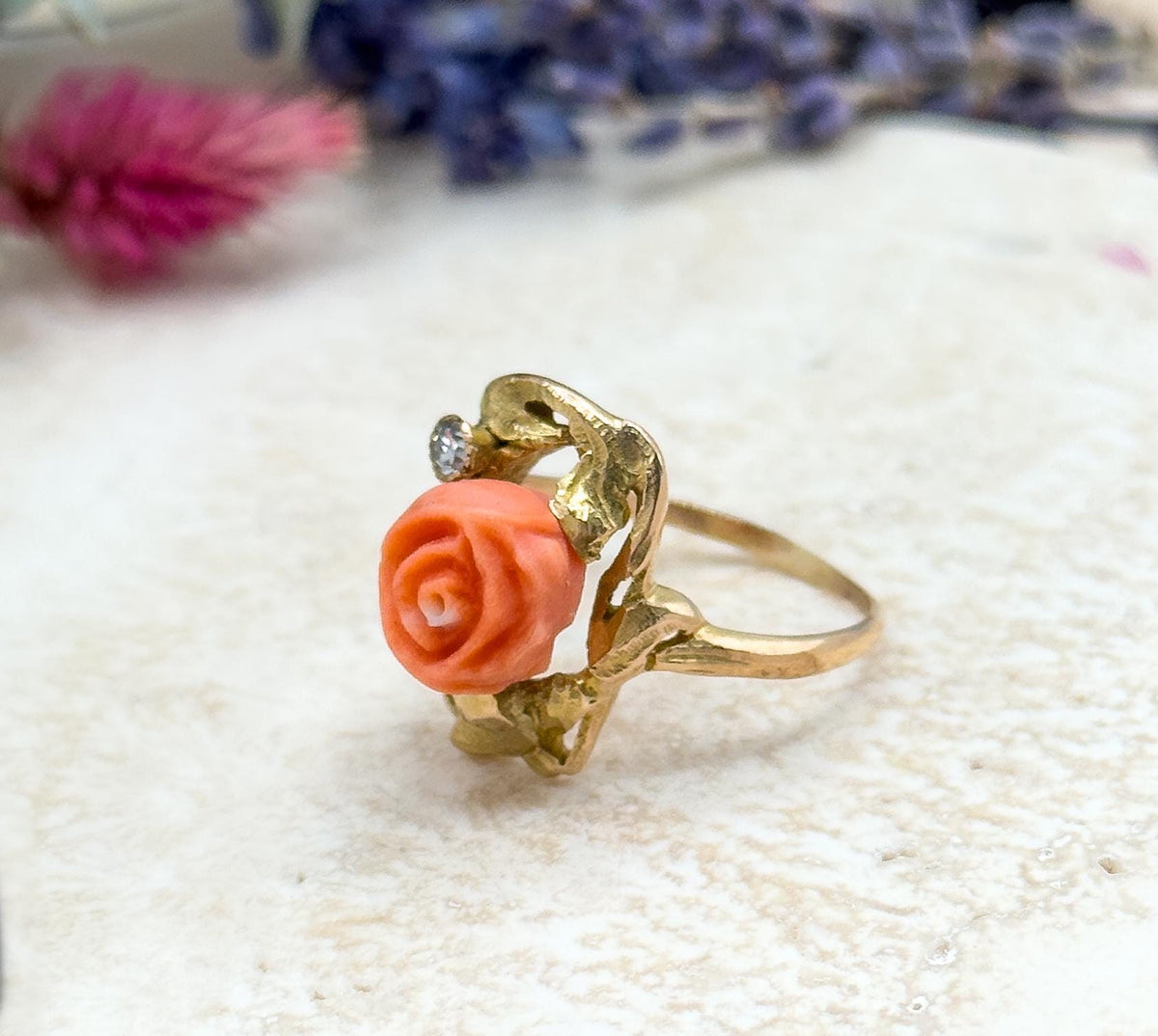 Angled three-quarter view of the ring standing upright on pale stone with dried pink and purple flowers blurred behind, showing the coral rose, bezel-set diamond and the slim gold shank from the side.