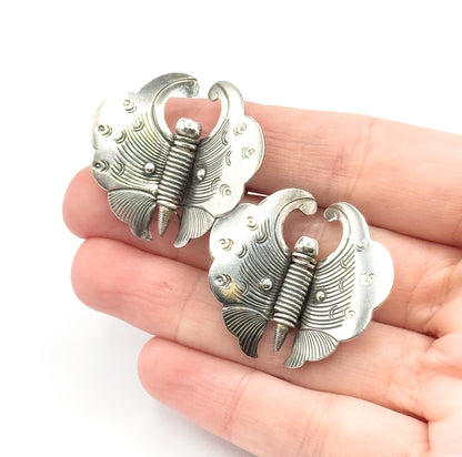 Two vintage sterling silver butterfly pins shown resting on open fingers for scale, their etched wings framing a ribbed body with patina settled into the recessed lines.
